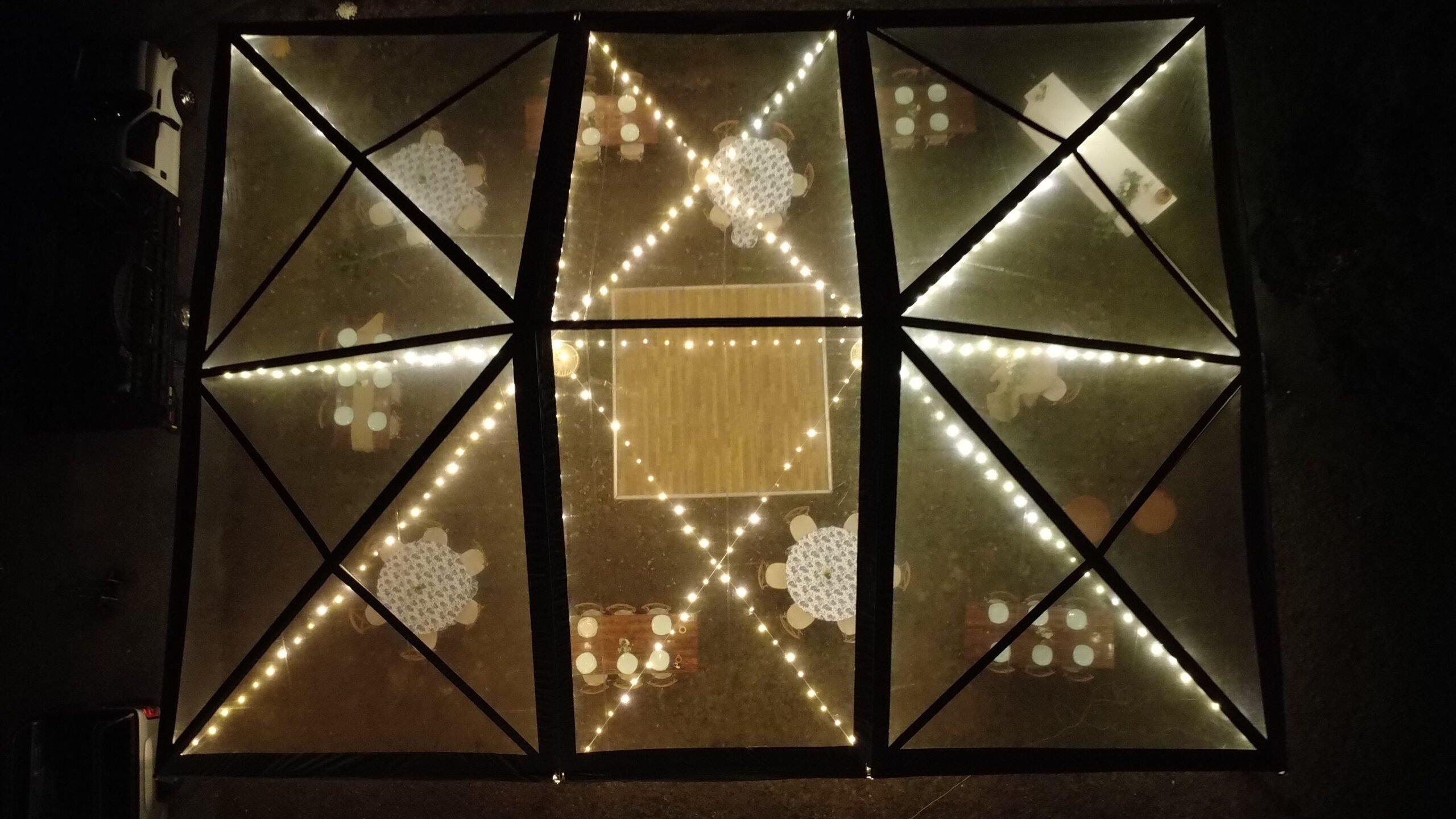 Aerial view of a tent illuminated with string lights, featuring elegantly arranged tables with decorative tablecloths, suitable for events like weddings and parties.