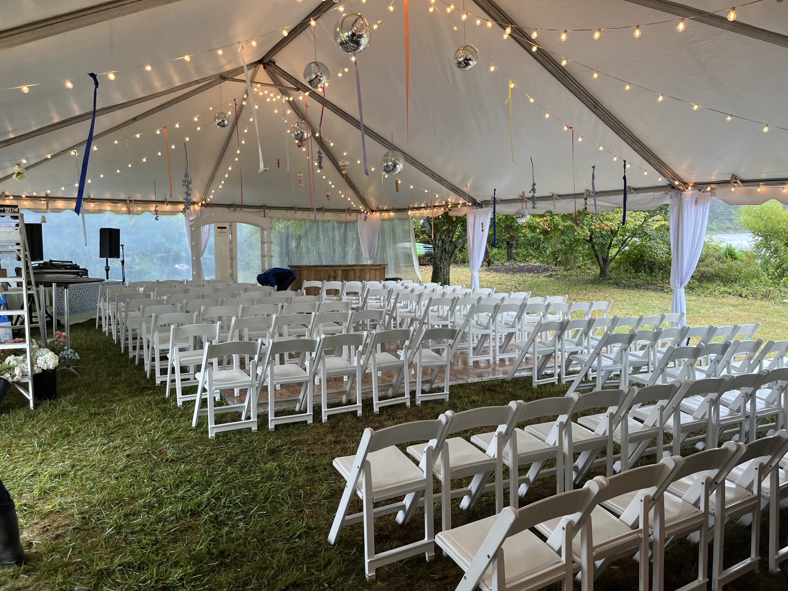 Tent interior decorated with string lights, white folding chairs arranged for an event, and colorful ribbons hanging from the ceiling, creating a charming atmosphere for weddings and celebrations.